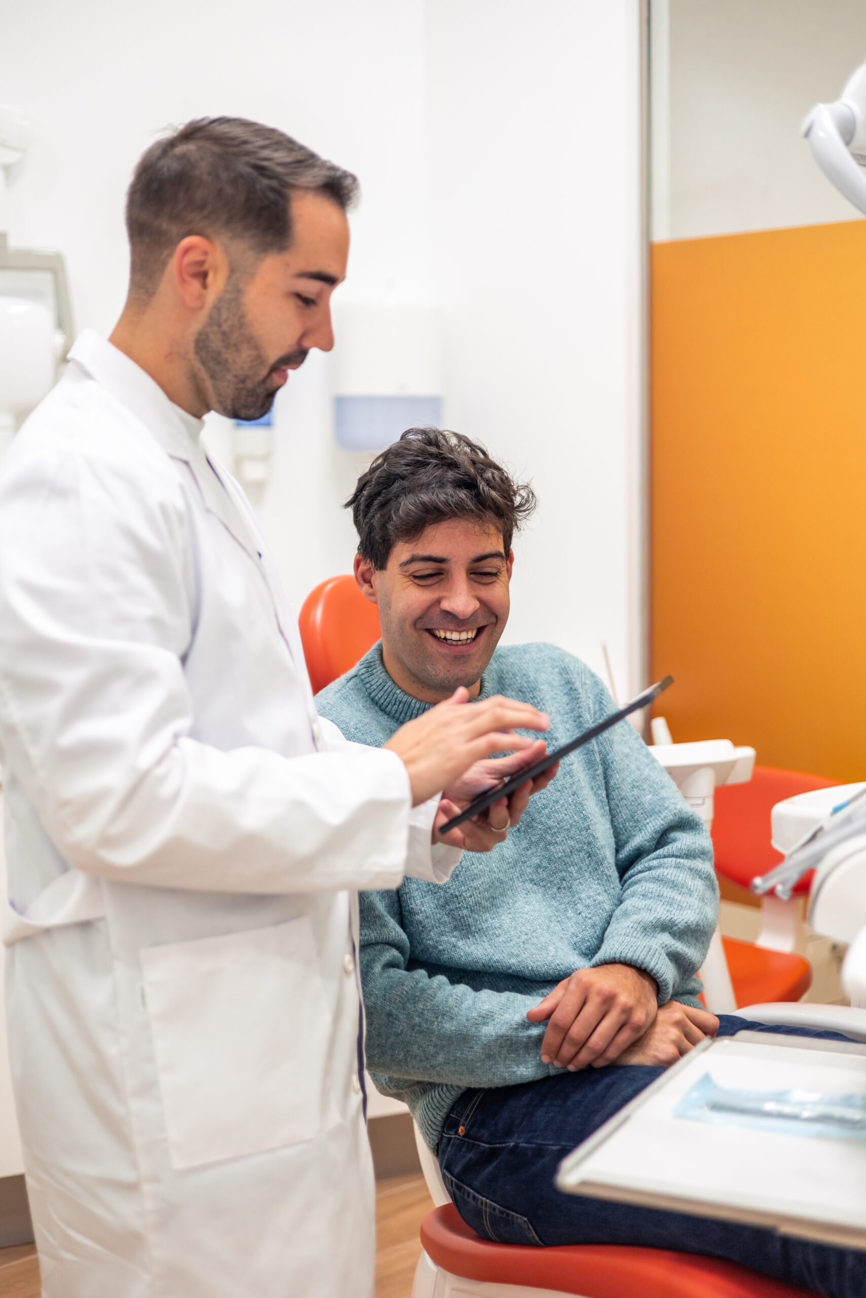 Dentist is holding a tablet and showing information to a smiling patient sitting in a dental chair during a consultation in a modern dental clinic
